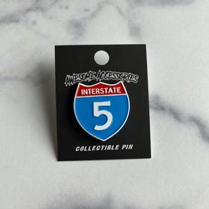 May include: A black and white collectible pin featuring a blue interstate highway sign with the number 5 in white. The sign has a red border and the words "Interstate" in white.