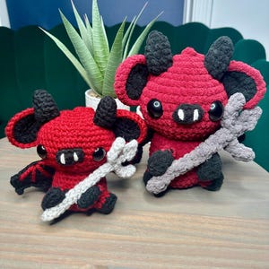 May include: Two handmade crochet devil plushies. Each plushie is red with black accents on the ears, wings, and horns. They have large black eyes and hold a gray trident. The plushies are sitting on a wooden surface.