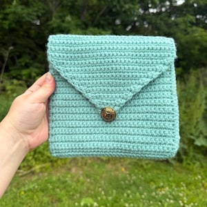 May include: A light mint green crochet envelope-style pouch featuring a textured stitch pattern and a triangular flap secured by a single round, antique-style bronze-toned metal button with an embossed decorative pattern on the front surface.