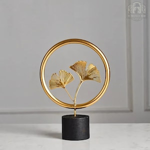 Puede incluir: Escultura de metal dorado de dos hojas de ginkgo dentro de un marco circular, sobre una base negra.