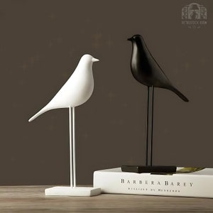 Könnte beinhalten: Zwei dekorative Vogelskulpturen, eine weiße und eine schwarze, stehen auf einer Holzoberfläche. Der weiße Vogel befindet sich links und der schwarze Vogel rechts. Beide Vögel haben lange, dünne Beine und stehen auf kleinen quadratischen Sockeln. Das Buch im Hintergrund trägt den Titel "BARBERA BAREY REALIZED BY HENREDON".