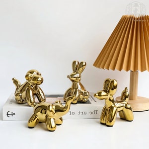 May include: Four gold-colored ceramic animal figurines in the shape of a rabbit, elephant, dog, and monkey. The figurines are sitting on a book with the text "The art of" visible. The figurines are all in a playful, balloon-like style.