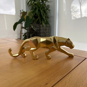 May include: A gold geometric panther figurine. The panther is in a crouching position with its head turned to the side. The figurine is made of a shiny metal and has a modern design.