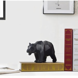 Puede incluir: Una escultura de oso geom&eacute;trico negro se encuentra sobre un libro dorado, con libros rojos y blancos detr&aacute;s.