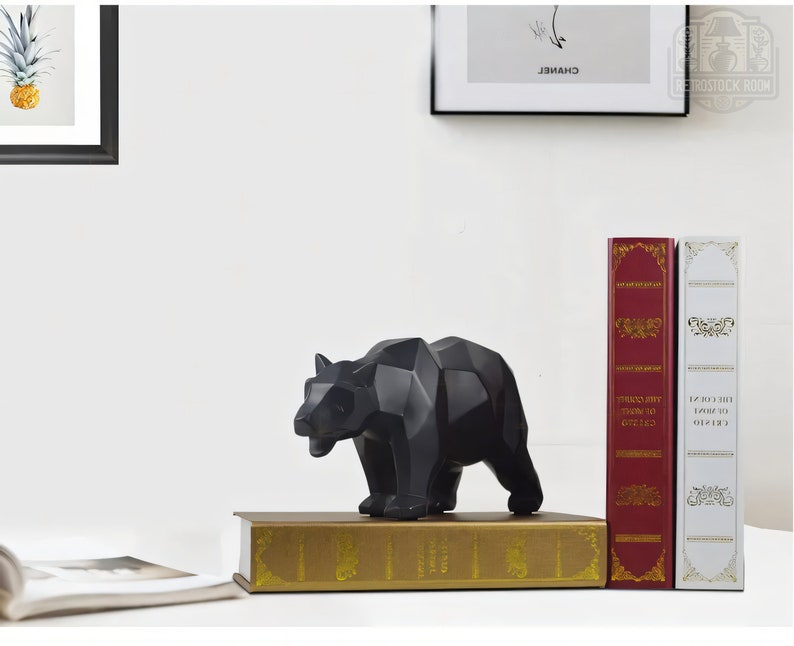 Puede incluir: Una escultura de oso geom&eacute;trico negro se encuentra sobre un libro dorado, con libros rojos y blancos detr&aacute;s.