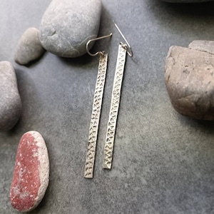 Peut inclure: Une paire de boucles d'oreilles en argent en forme de barres avec un motif géométrique. Les boucles d'oreilles rectangulaires sont suspendues à un crochet. Elles sont présentées sur une surface grise avec des pierres grises et roses.
