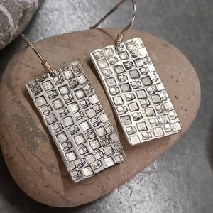 Könnte beinhalten: Ein Paar silberne Ohrringe mit geometrischem Muster. Die Ohrringe sind rechteckig und haben eine strukturierte Oberfläche.