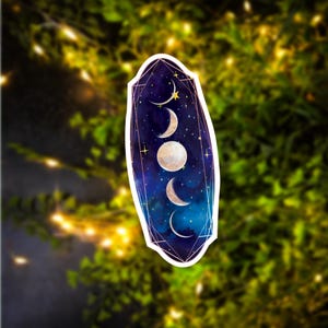 May include: A sticker with a celestial design featuring a crescent moon, full moon, and stars on a dark blue background with a gold geometric frame.