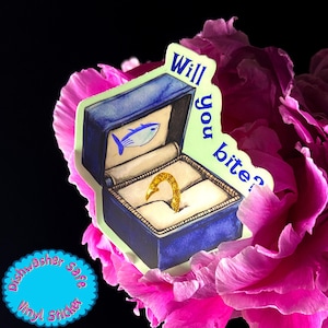 May include: A vinyl sticker featuring a blue ring box with a gold hook-shaped ring inside. The box has a fish illustration and the words "Will you bite?" The sticker is surrounded by pink flower petals.