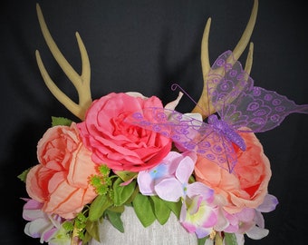 May include: A floral headpiece featuring faux antlers, coral and pink roses, and a purple glitter butterfly. The arrangement includes peach and pink flowers, green leaves, and white cascading blossoms, all set on a neutral-toned head form.