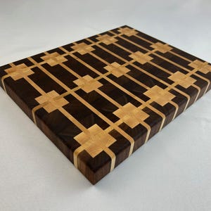 Könnte beinhalten: Rechteckiges Schneidebrett mit geometrischem Muster. Das Brett besteht aus dunkelbraunem und hellbraunem Holz mit einem sich wiederholenden Design aus Quadraten und Linien. Das Brett ist ca. 30 cm lang und 23 cm breit.