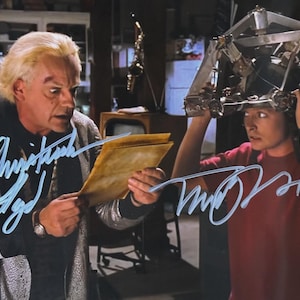 May include: A photograph featuring two men, one holding a document and the other holding a metallic headpiece. The man on the left wears a silver jacket and the man on the right wears a red shirt. Both have signatures in blue ink.