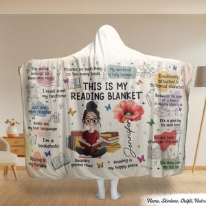 Op de afbeelding: Een witte leesdeken met capuchon met de tekst "THIS IS MY READING BLANKET" en illustraties van boeken, vlinders en een lezende vrouw. De deken bevat citaten over lezen.