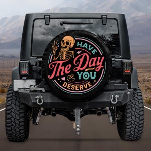 Puede incluir: Jeep negro con una funda para la rueda de repuesto. La funda muestra un esqueleto haciendo el signo de la paz, con el texto "Have The Day You Deserve" en rosa, turquesa y blanco sobre fondo negro.