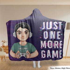 Puede incluir: Una manta con capucha con una ilustración de dibujos animados de una persona con auriculares y sosteniendo un mando de juego. La manta tiene el texto "JUST ONE MORE GAME" en blanco y morado. El nombre de la persona, "WILLIAM", está en su camiseta.