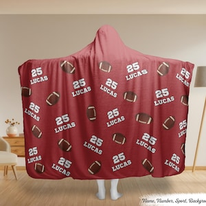 Op de afbeelding: Een bordeauxrode capuchondeken met een herhalend patroon van bruine American footballs en de witte tekst "25 LUCAS". De deken is ontworpen om te worden gedragen en biedt warmte en comfort.