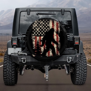 May include: Black spare wheel cover with a silhouette of Bigfoot walking in front of a distressed American flag. The flag design includes red and white stripes and white stars. The cover is mounted on a black Jeep.