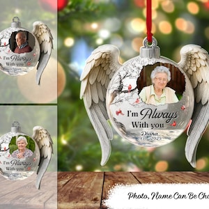 Puede incluir: Un adorno conmemorativo personalizado con alas de ángel. El adorno de cristal transparente presenta una foto y el texto "I'm Always With You". El adorno está suspendido por una cinta roja. El texto "Photo, Name Can Be Changed" está en la parte inferior.