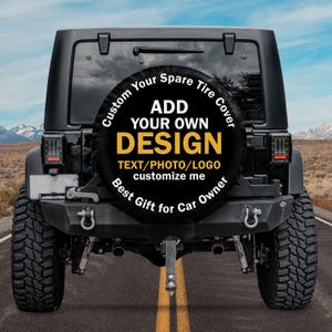 Könnte beinhalten: Schwarzer Jeep mit einer individuellen Reserveradabdeckung. Die Abdeckung ist schwarz mit weißem und gelbem Text, der "ADD YOUR OWN DESIGN" und "Best Gift for Car Owner" lautet. Der Hintergrund zeigt eine Straße und Berge.
