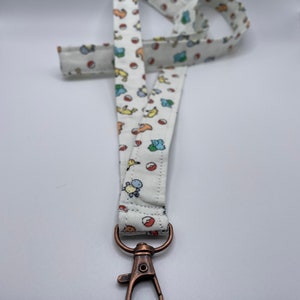 May include: A white fabric lanyard featuring a repeating pattern of small, colorful cartoon creatures and red and white spherical icons. The strap is attached to a metallic, copper-toned swivel lobster clasp hook at the bottom end.