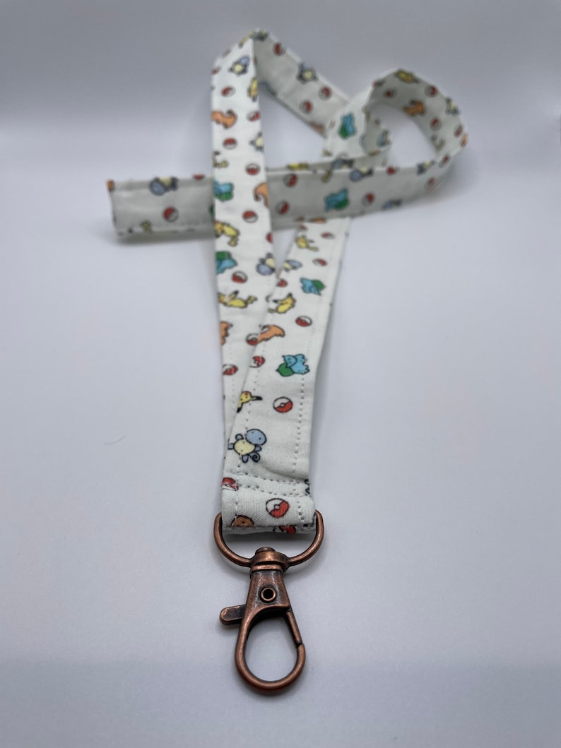 May include: A white fabric lanyard featuring a repeating pattern of small, colorful cartoon creatures and red and white spherical icons. The strap is attached to a metallic, copper-toned swivel lobster clasp hook at the bottom end.