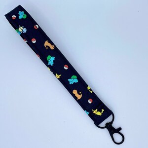 May include: A fabric wristlet keychain featuring a dark navy blue background with a repeating pattern of colorful cartoon creatures and red and white circular icons. The strap is finished with a black metal lobster clasp at one end.