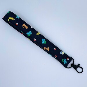May include: A black fabric wristlet keychain featuring a repeating pattern of colorful cartoon creatures and red and white spherical icons. The strap is secured with a black metal lobster clasp attached to one end of the folded fabric loop.