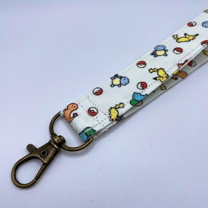 May include: A white fabric wrist lanyard featuring a repeating pattern of small colorful cartoon creatures and red and white circular balls. The fabric strap is attached to a circular metal ring connected to a bronze-toned metal lobster clasp.