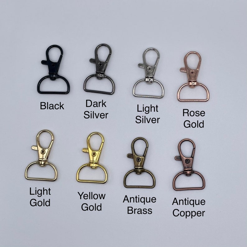 May include: Eight metal lobster claw clasps with D-rings in various finishes: black, dark silver, light silver, rose gold, light gold, yellow gold, antique brass, and antique copper. Each clasp is labeled with its corresponding color name below it.