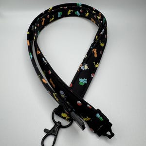 May include: A black fabric lanyard featuring a repeating pattern of colorful small cartoon creatures and red and white spherical icons. The lanyard includes a black metal lobster clasp and a black plastic buckle attachment at the bottom end.