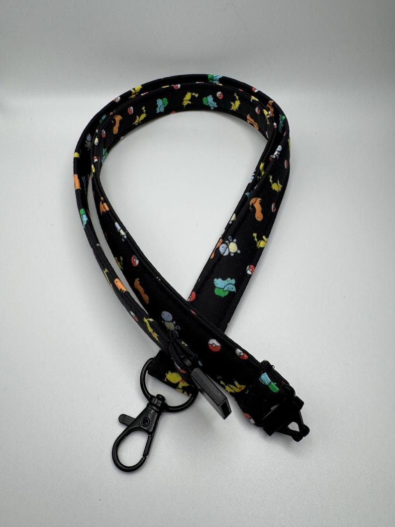 May include: A black fabric lanyard featuring a repeating pattern of colorful small cartoon creatures and red and white spherical icons. The lanyard includes a black metal lobster clasp and a black plastic buckle attachment at the bottom end.