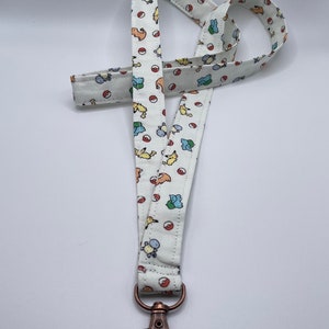 May include: A white fabric lanyard featuring a repeating pattern of small colorful cartoon creatures and red and white spherical icons. The strap is finished with a metallic bronze-toned lobster clasp attached to the bottom end of the fabric loop.