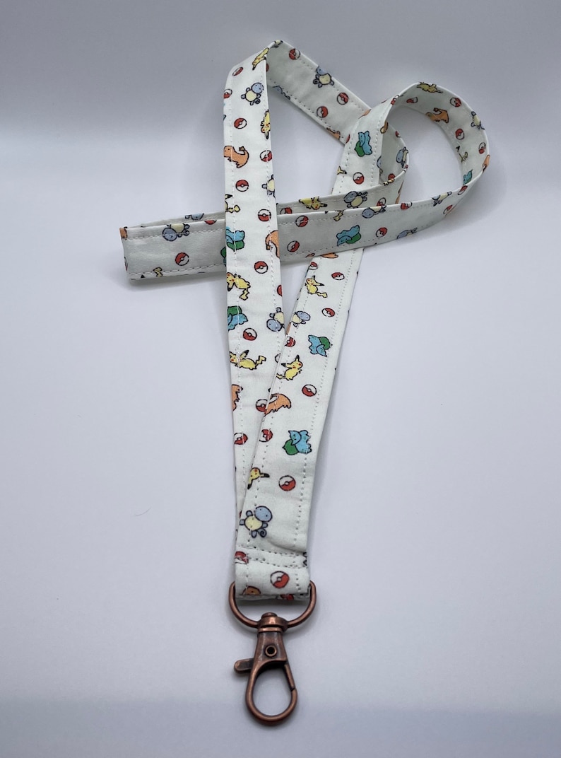 May include: A white fabric lanyard featuring a repeating pattern of small colorful cartoon creatures and red and white spherical icons. The strap is finished with a metallic bronze-toned lobster clasp attached to the bottom end of the fabric loop.