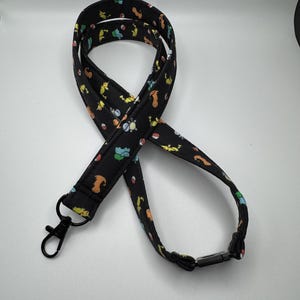 May include: A black fabric lanyard featuring a repeating pattern of colorful cartoon creatures and red and white spheres. The lanyard includes a black metal lobster clasp at one end and a black plastic breakaway buckle at the back of the neck strap.