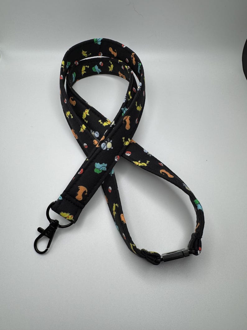 May include: A black fabric lanyard featuring a repeating pattern of colorful cartoon creatures and red and white spheres. The lanyard includes a black metal lobster clasp at one end and a black plastic breakaway buckle at the back of the neck strap.
