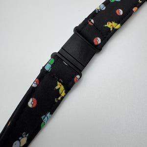 May include: A black fabric strap featuring a repeating pattern of small, colorful cartoon characters and red and white spheres. The strap includes a black plastic side-release buckle in the center, allowing for adjustable length and secure fastening.