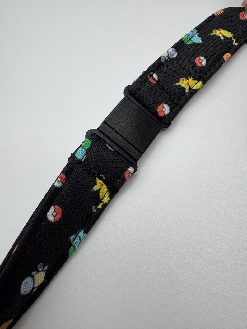 May include: A black fabric strap featuring a repeating pattern of small, colorful cartoon characters and red and white spheres. The strap includes a black plastic side-release buckle in the center, allowing for adjustable length and secure fastening.