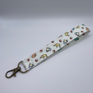 May include: A white fabric wristlet keychain featuring a repeating pattern of small yellow, blue, and orange cartoon creatures alongside red and white circular balls. The strap is attached to a circular ring with an antique brass-toned metal lobster clasp.