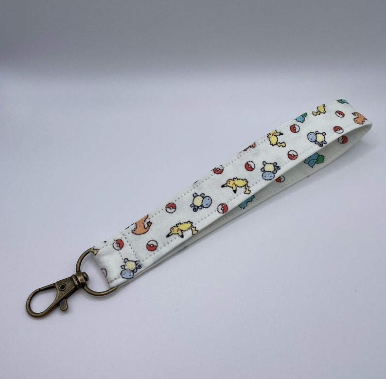 May include: A white fabric wristlet keychain featuring a repeating pattern of small yellow, blue, and orange cartoon creatures alongside red and white circular balls. The strap is attached to a circular ring with an antique brass-toned metal lobster clasp.