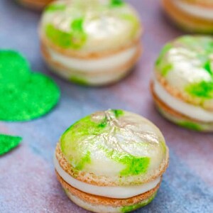 K&ouml;nnte beinhalten: Gr&uuml;ne und goldene Macarons mit wei&szlig;em Frosting. Die Macarons sind auf einer rosa und goldenen Oberfl&auml;che angeordnet.