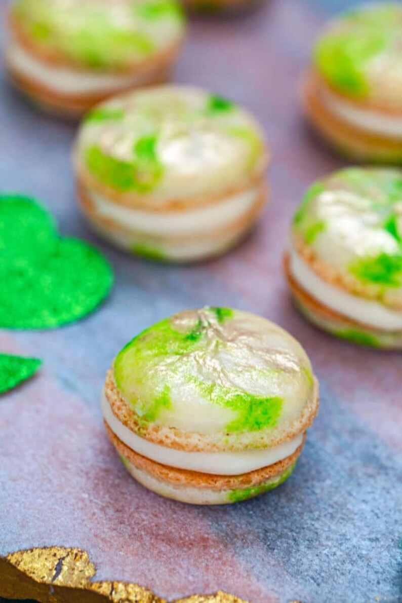 K&ouml;nnte beinhalten: Gr&uuml;ne und goldene Macarons mit wei&szlig;em Frosting. Die Macarons sind auf einer rosa und goldenen Oberfl&auml;che angeordnet.
