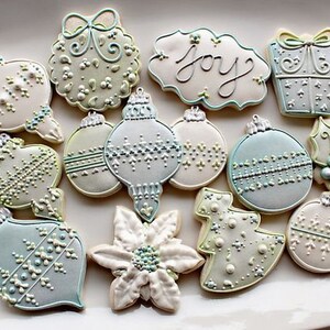 K&ouml;nnte beinhalten: Eine Sammlung von 12 weihnachtlichen Zuckerpl&auml;tzchen, die mit wei&szlig;em, blauem und gr&uuml;nem Zuckerguss verziert sind. Die Pl&auml;tzchen sind in Form von Kugeln, einem Weihnachtsbaum, einer Weihnachtsstern und einer Geschenkbox geformt. Ein Pl&auml;tzchen tr&auml;gt die Aufschrift "Joy".