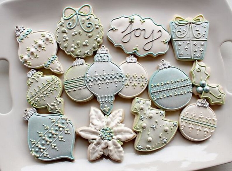 K&ouml;nnte beinhalten: Eine Sammlung von 12 weihnachtlichen Zuckerpl&auml;tzchen, die mit wei&szlig;em, blauem und gr&uuml;nem Zuckerguss verziert sind. Die Pl&auml;tzchen sind in Form von Kugeln, einem Weihnachtsbaum, einer Weihnachtsstern und einer Geschenkbox geformt. Ein Pl&auml;tzchen tr&auml;gt die Aufschrift "Joy".
