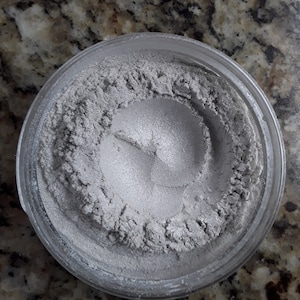 Puede incluir: Un pequeño frasco de vidrio lleno de polvo plateado brillante. El polvo es suave y tiene un ligero brillo.