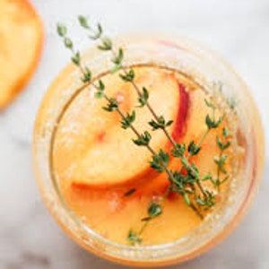May include: A glass filled with a peach smoothie topped with fresh peach slices and sprigs of thyme.