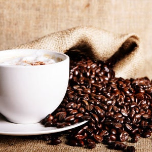 May include: A white ceramic coffee mug filled with coffee and a layer of foam on top. The mug is sitting on a white saucer. A burlap sack filled with coffee beans is in the background.