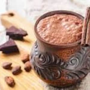 Könnte beinhalten: Eine braune Keramik-Tasse gefüllt mit einem schaumigen, schokoladenfarbenen Getränk. Zwei Zimtstangen liegen an der Seite der Tasse. Kakaobohnen und Stücke dunkler Schokolade sind um die Tasse herum auf einem hölzernen Schneidebrett verstreut.