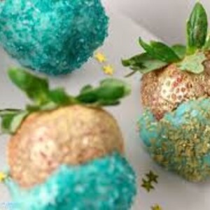 K&ouml;nnte beinhalten: Drei Erdbeeren, die in Schokolade getaucht und mit goldenen und t&uuml;rkisfarbenen Streuseln verziert sind. Die Erdbeeren sind auf einer wei&szlig;en Oberfl&auml;che mit goldenen Sternen angeordnet.