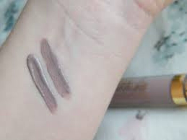 Puede incluir: Dos muestras de una sombra de ojos crema marr&oacute;n claro en la mu&ntilde;eca de una persona. La sombra de ojos est&aacute; en un tubo con una tapa dorada.