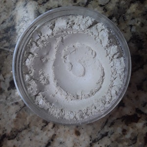 Puede incluir: Un pequeño frasco de vidrio transparente lleno de polvo gris claro. El polvo tiene un patrón de espiral.